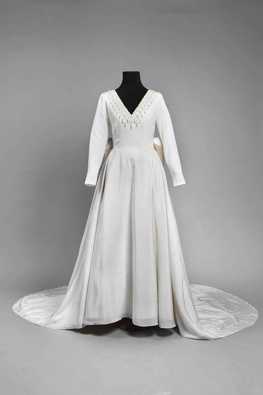 Se observa un maniquí negro llevando un vestido de novia blanco, de tafetán y raso, con manga larga, escote en V y cola